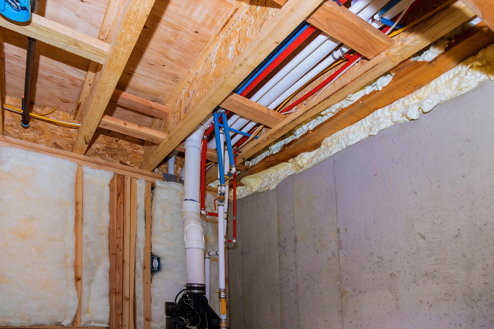 Exposed pipes, wiring, and insulation are visible along the ceiling and wall of an unfinished basement with concrete and wood framing.