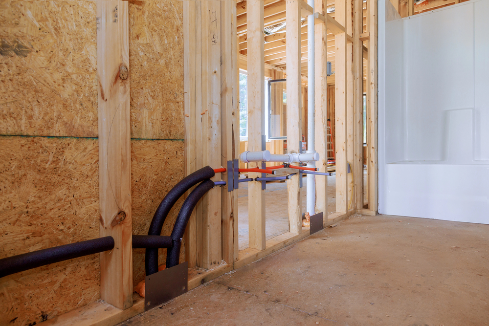 Exposed plumbing pipes and electrical wiring run through wooden wall framing in a building under construction, with a shower stall visible in the background.