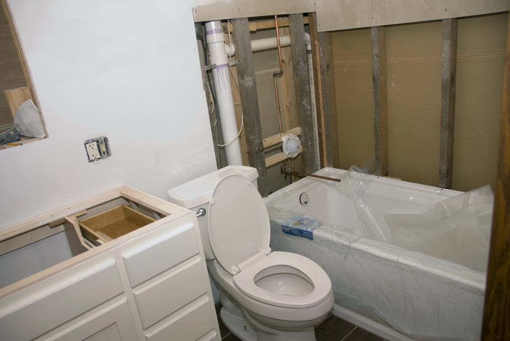 Partially completed bathroom under renovation, with exposed wall framing, installed toilet, uncovered bathtub, and unfinished vanity.