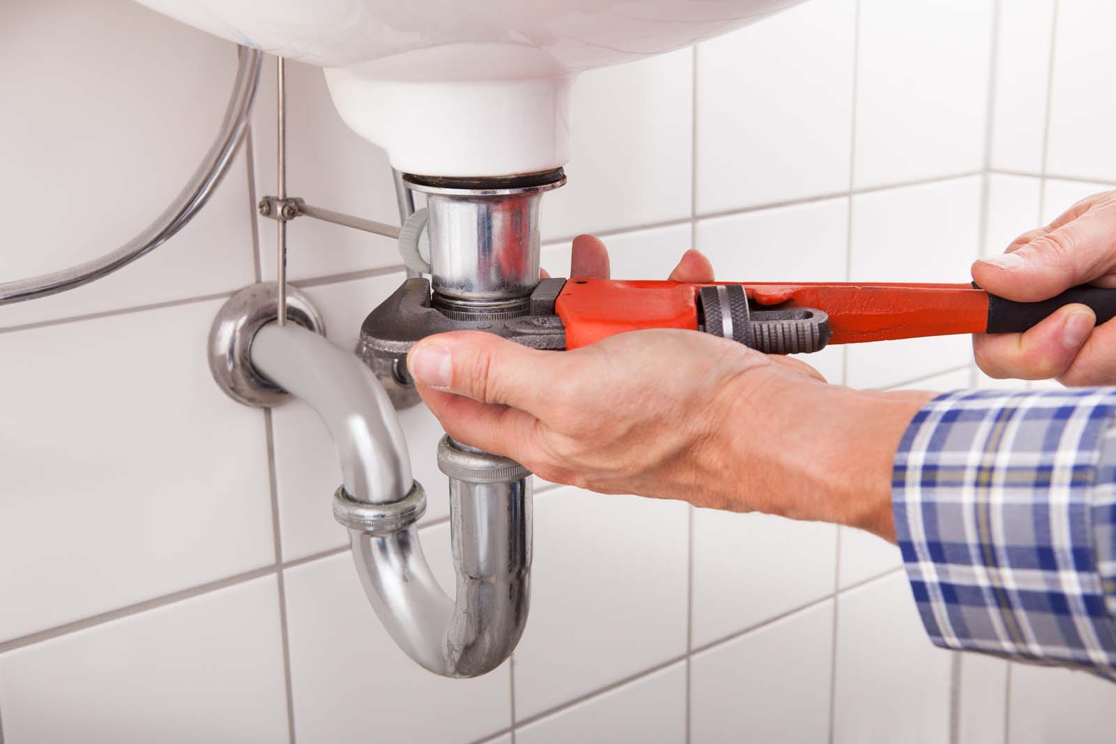 A plumber in a plaid shirt uses a pipe wrench to tighten or loosen a metal pipe joint under a white sink in a tiled bathroom.