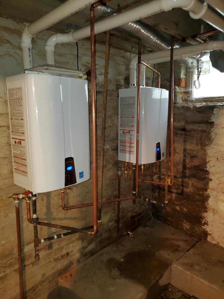 Two white tankless water heaters are mounted on a basement wall with exposed copper pipes and ventilation ducts visible.