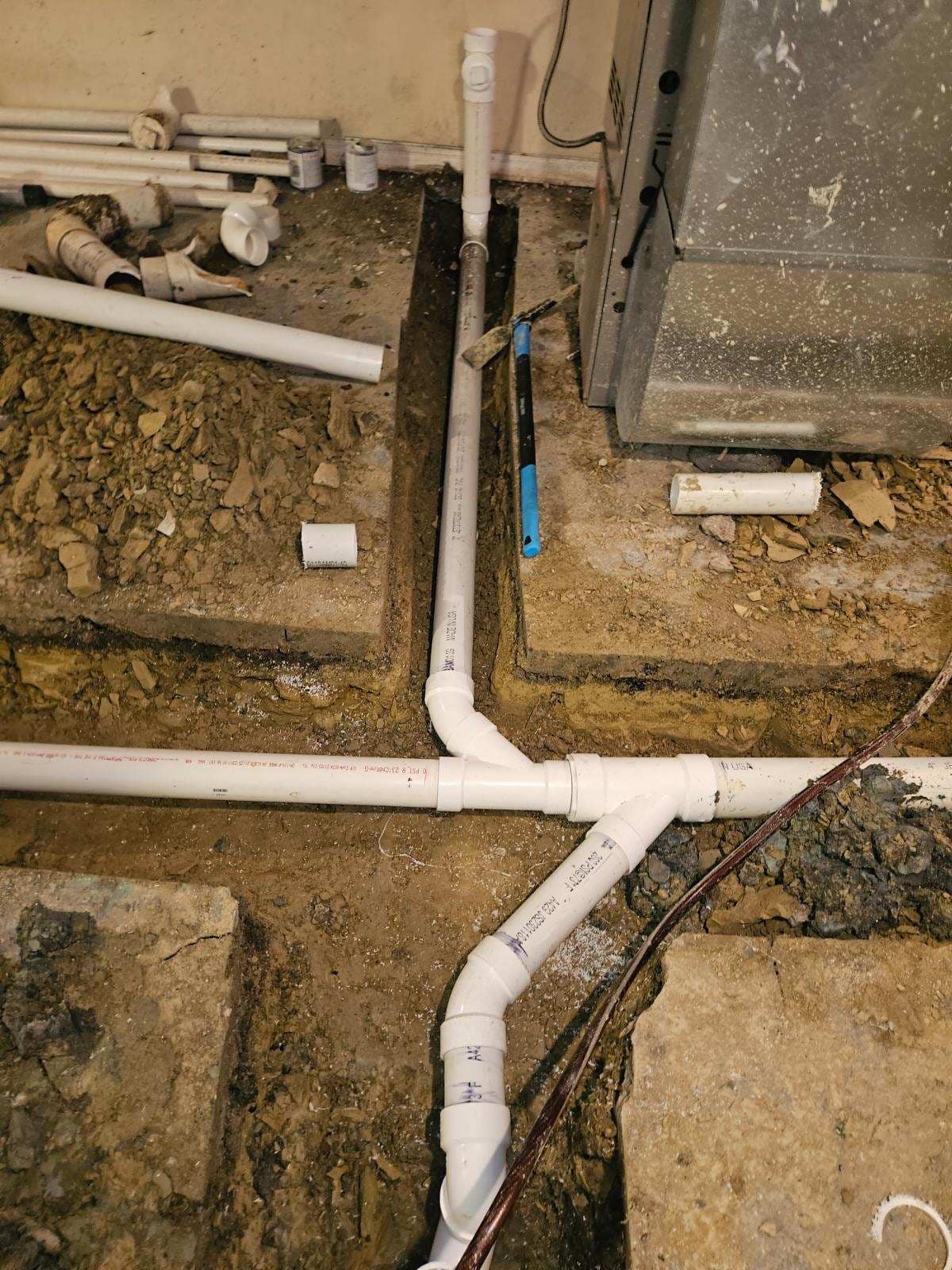 PVC pipes arranged and connected in trenches in a concrete floor, likely for plumbing installation or repair, with tools and pipe fittings visible nearby.