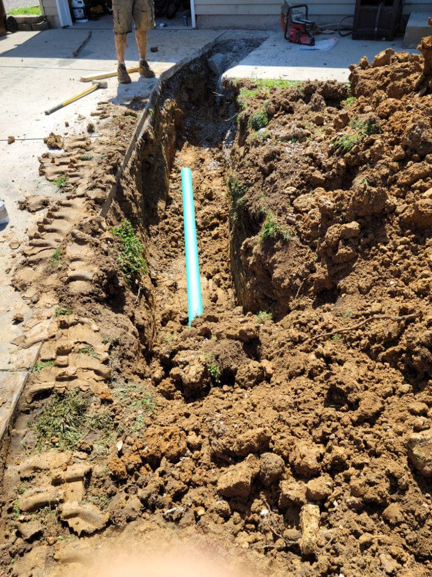 A trench dug alongside a driveway with a section of blue pipe placed inside, piles of dirt on both sides, and a person standing in the background.